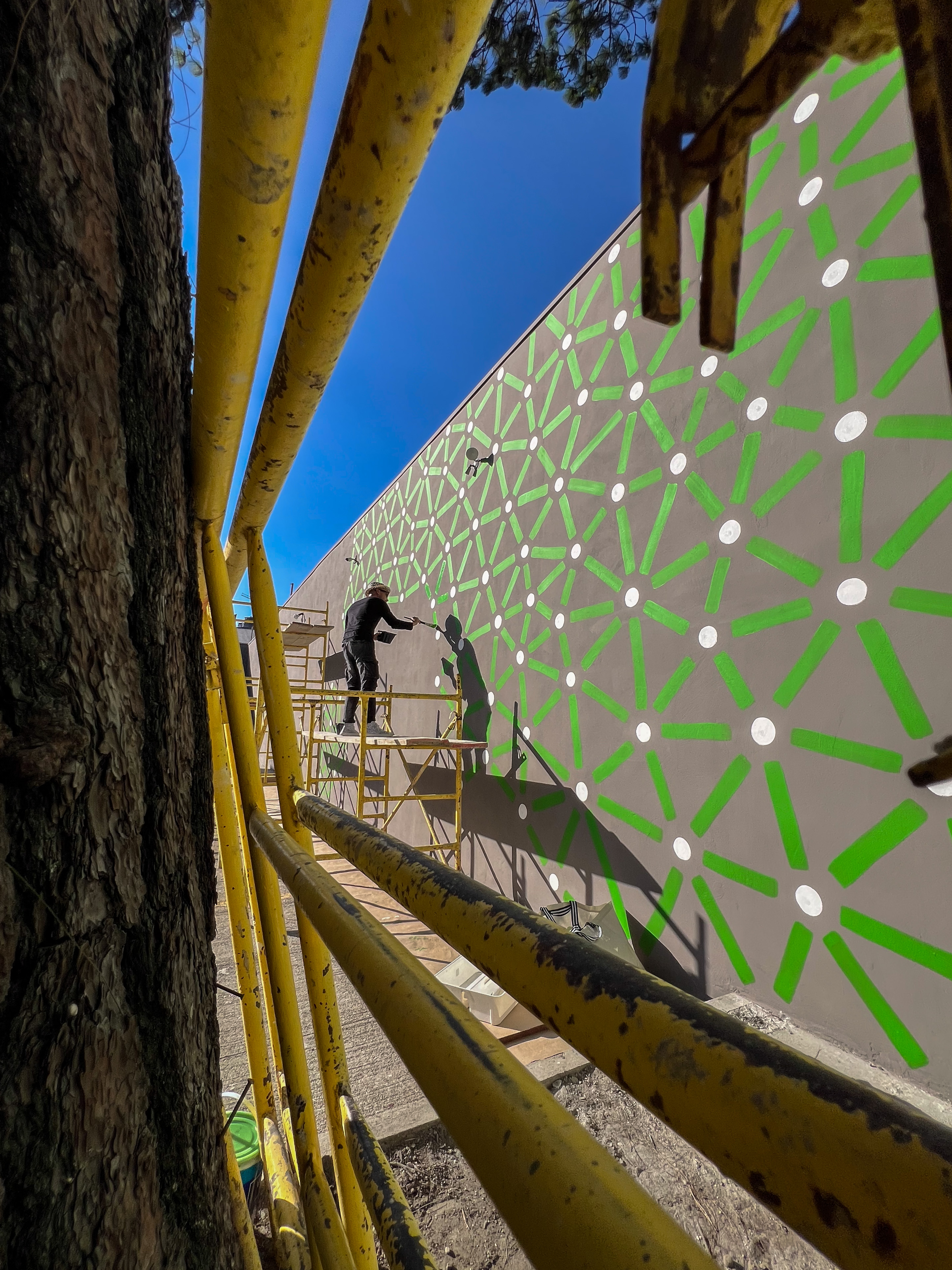 Roots: Artist works from yellow scaffolding to paint green lines and pale dots on a tall exterior wall.