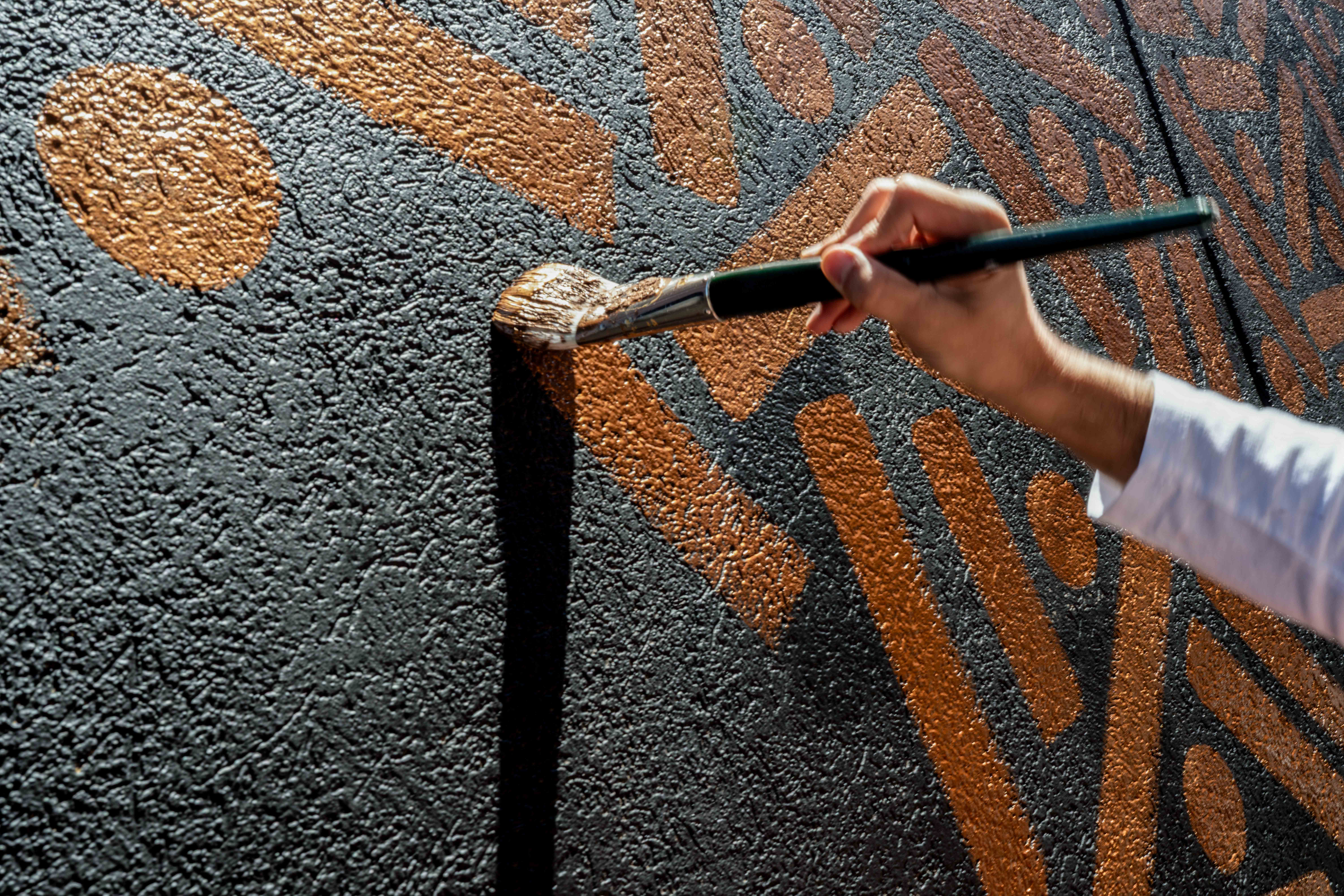 The Golden Weave: Close-up of an artist’s brush adding golden paint to a dark exterior wall.