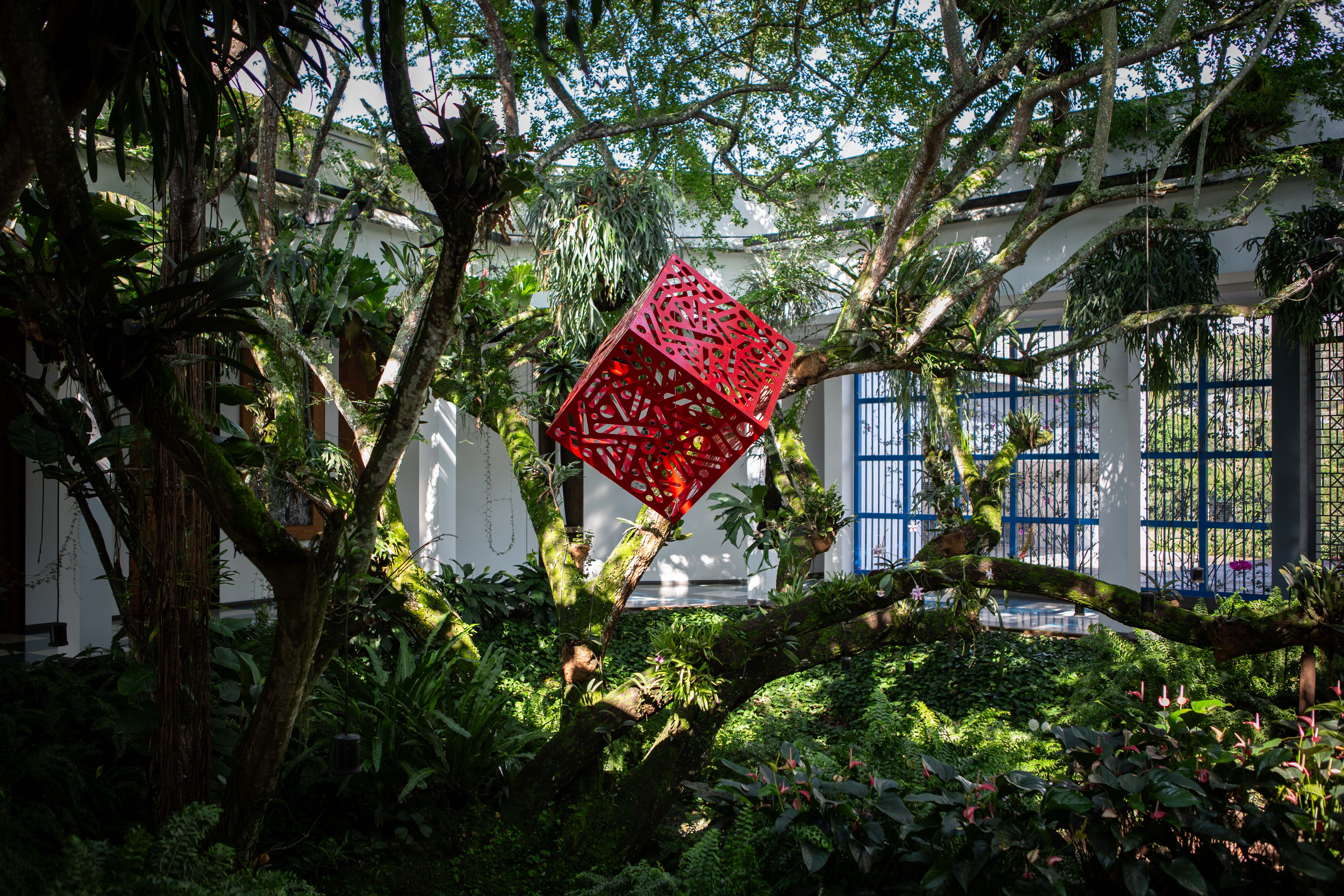Hemoglobin: Red cube sculpture with patterned cutouts suspended among trees in a garden.