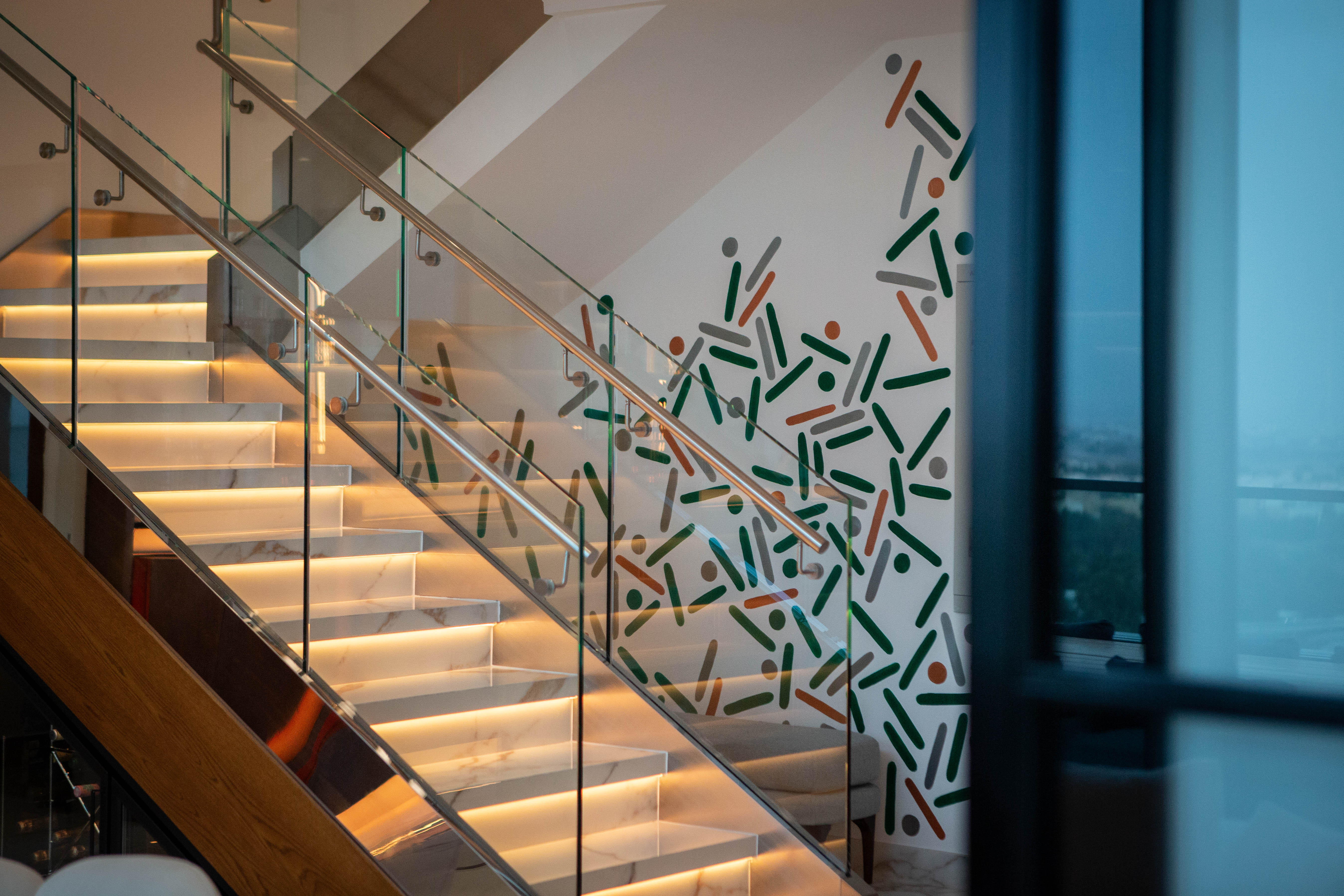 Green and dark dots and lines decorate a glass staircase railing beside illuminated wooden steps.