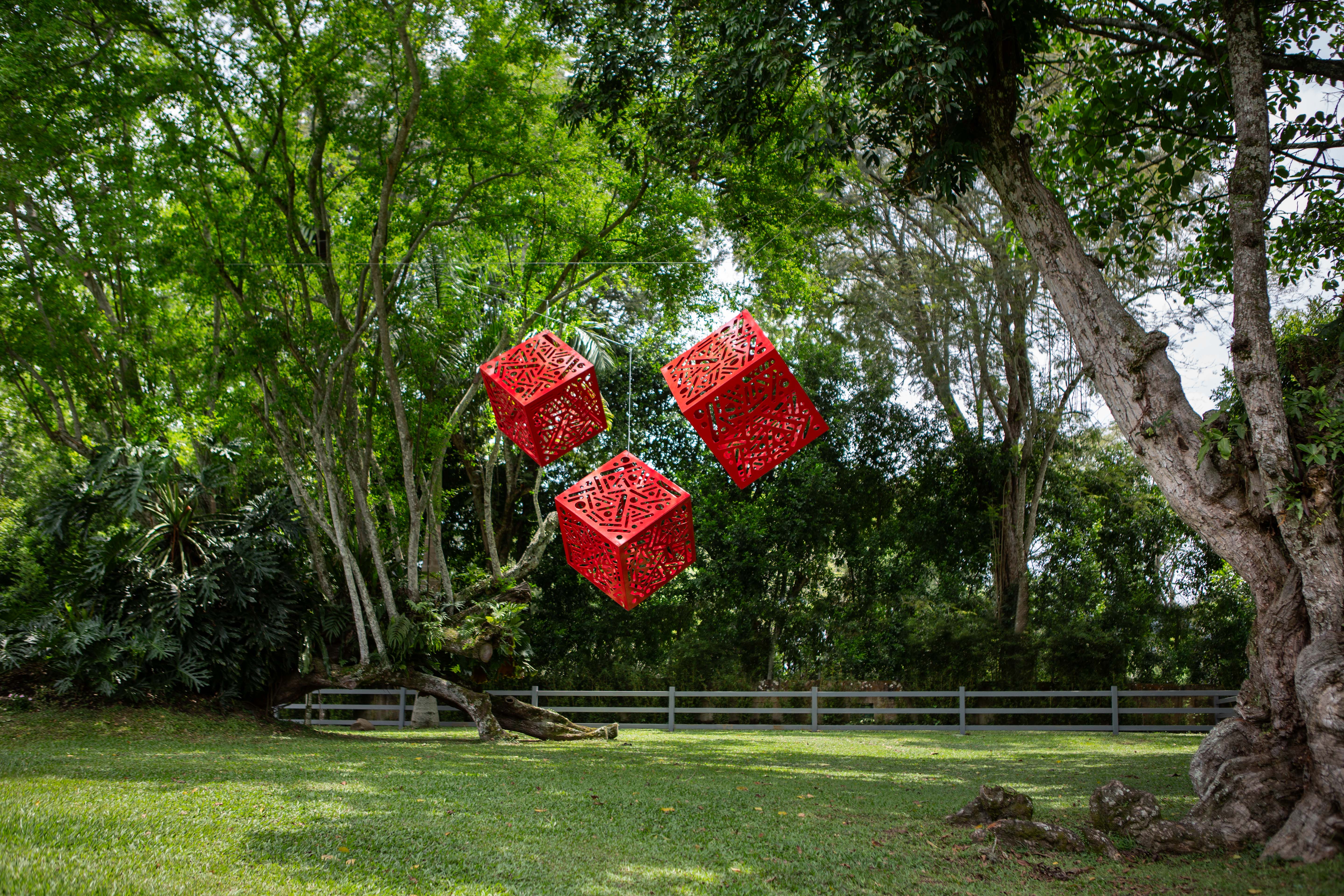 Hemoglobin: Three red cutout cube sculptures suspended among leafy garden trees.