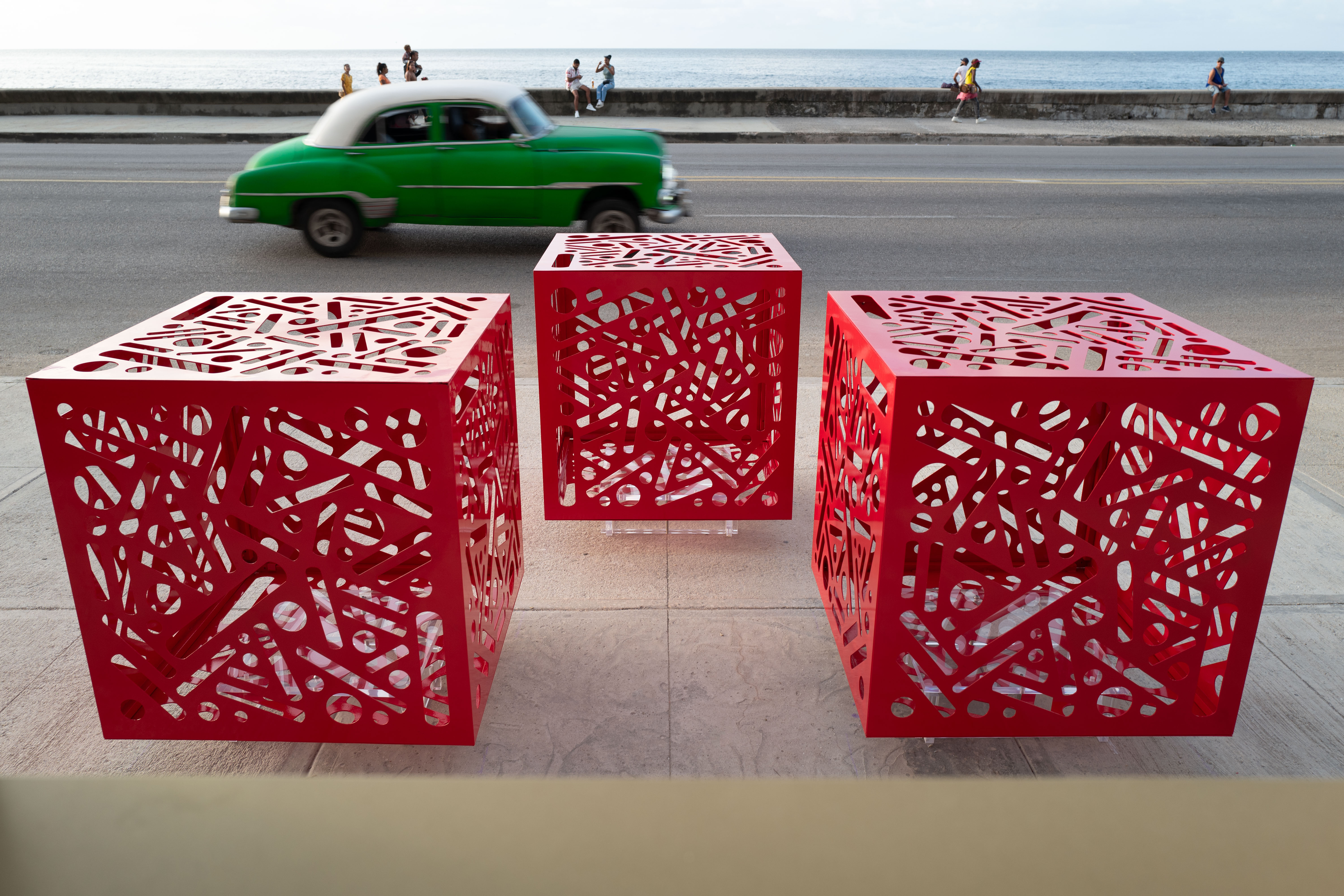 Three red cube sculptures with patterned cutouts displayed beside a waterfront road.