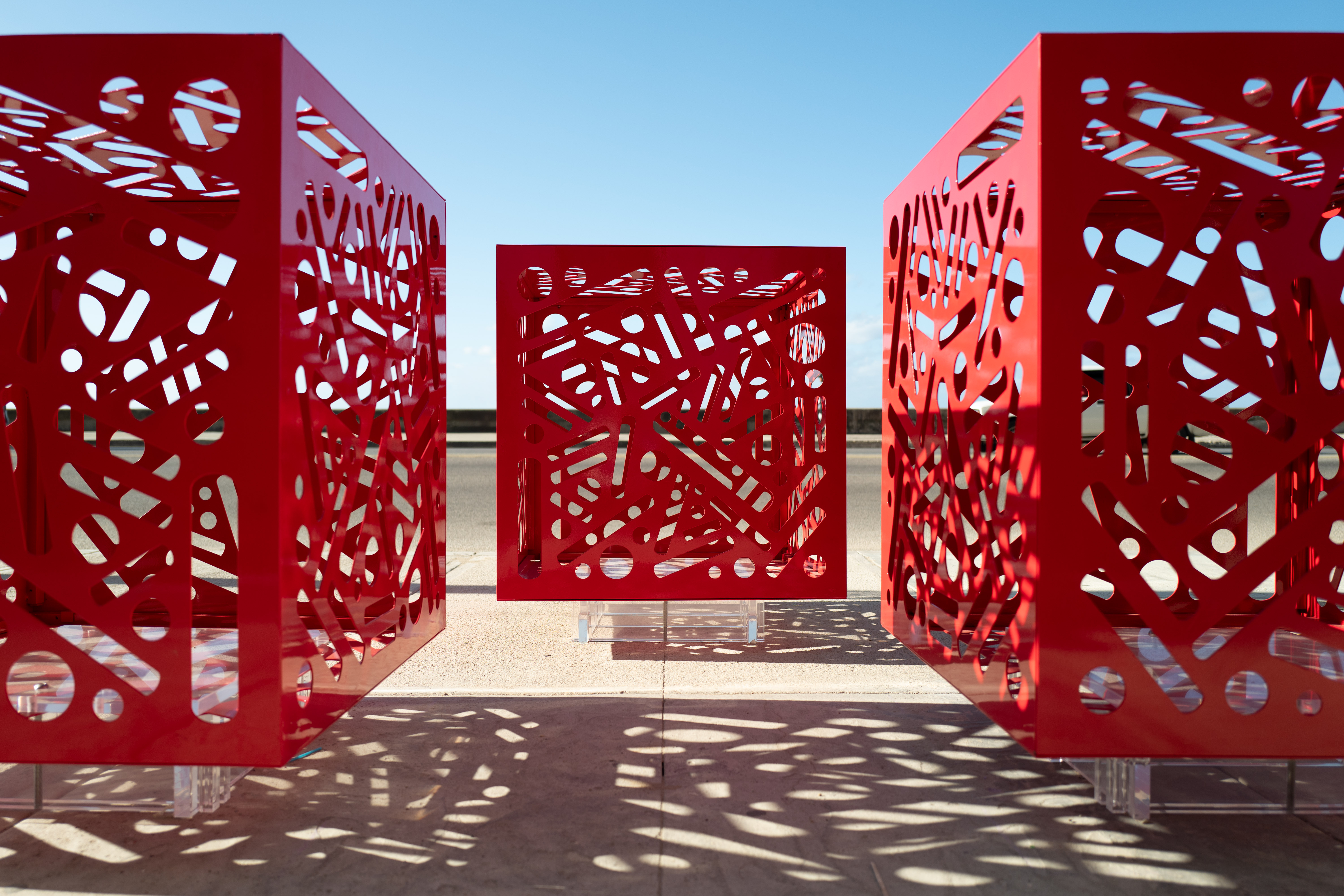 Hemoglobin - Installation of 3: Three red cutout cube sculptures cast geometric shadows onto sunlit pavement.