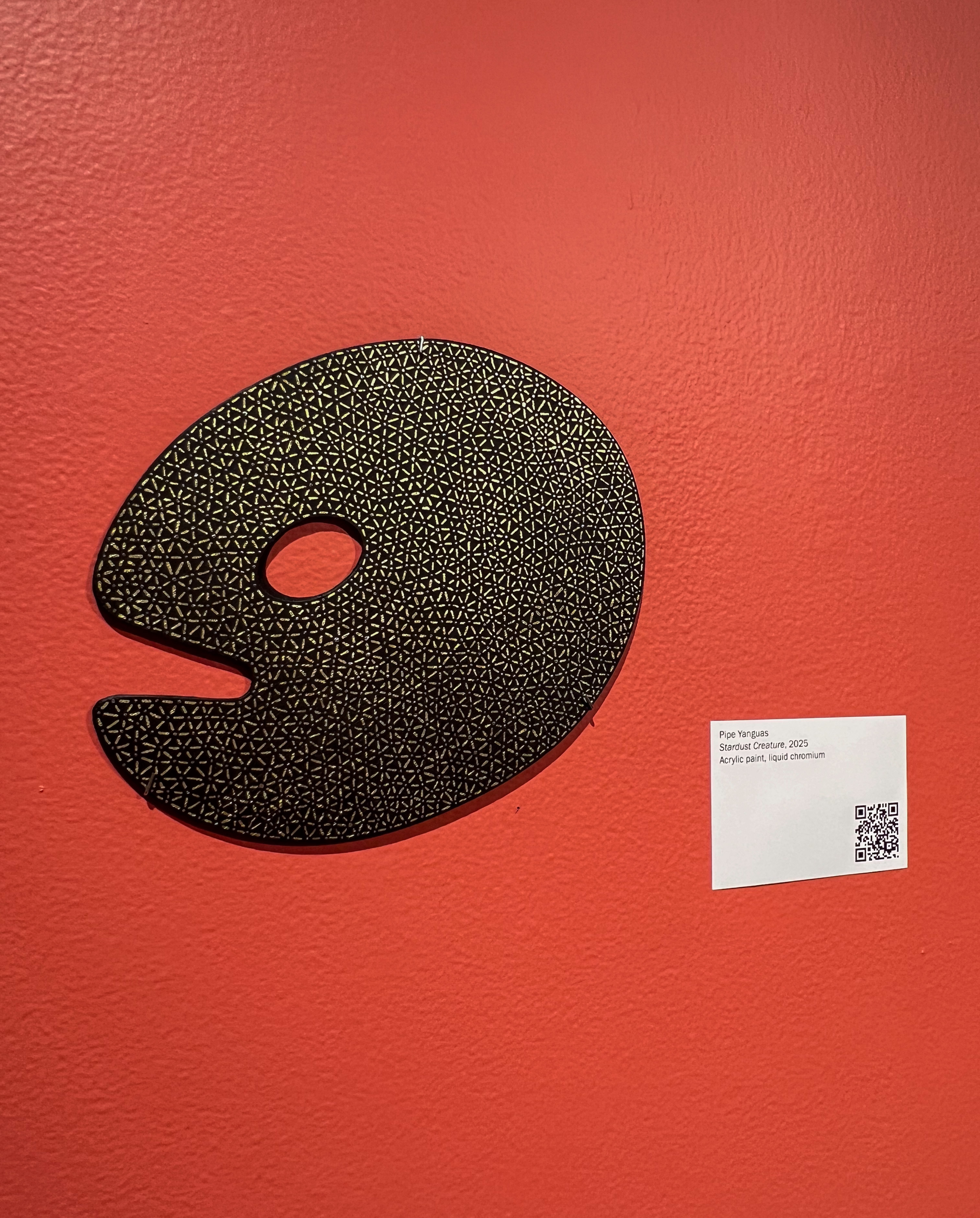 “Stardust Creature” (2026) by Pipe Yanguas, featuring a black background with intricate silver and gold dots and lines, displayed on a red wall at the Lowe Art Museum during The Palette Project.