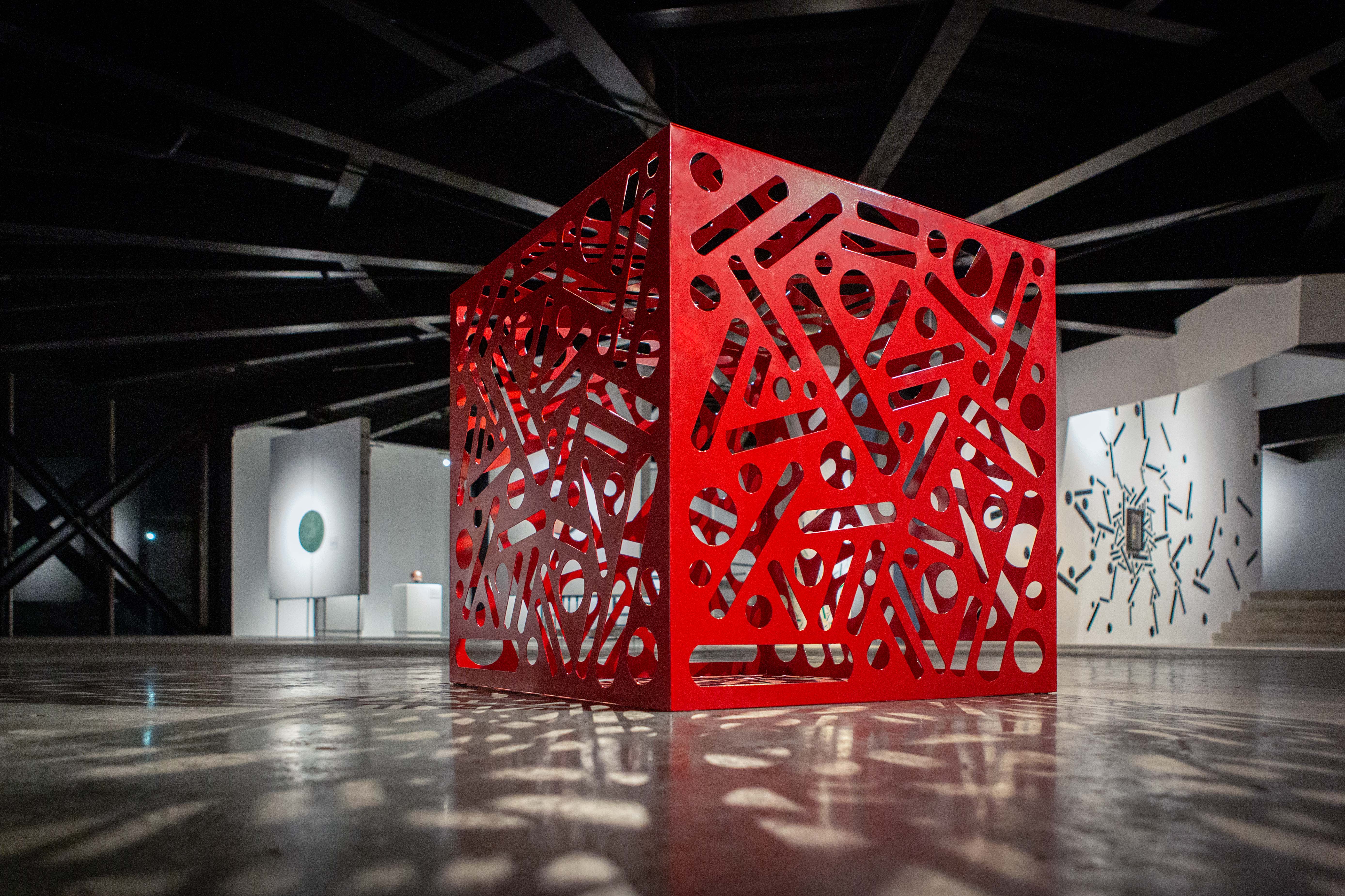 Red cube sculpture with cutout dots and lines stands in a dark exhibition gallery.