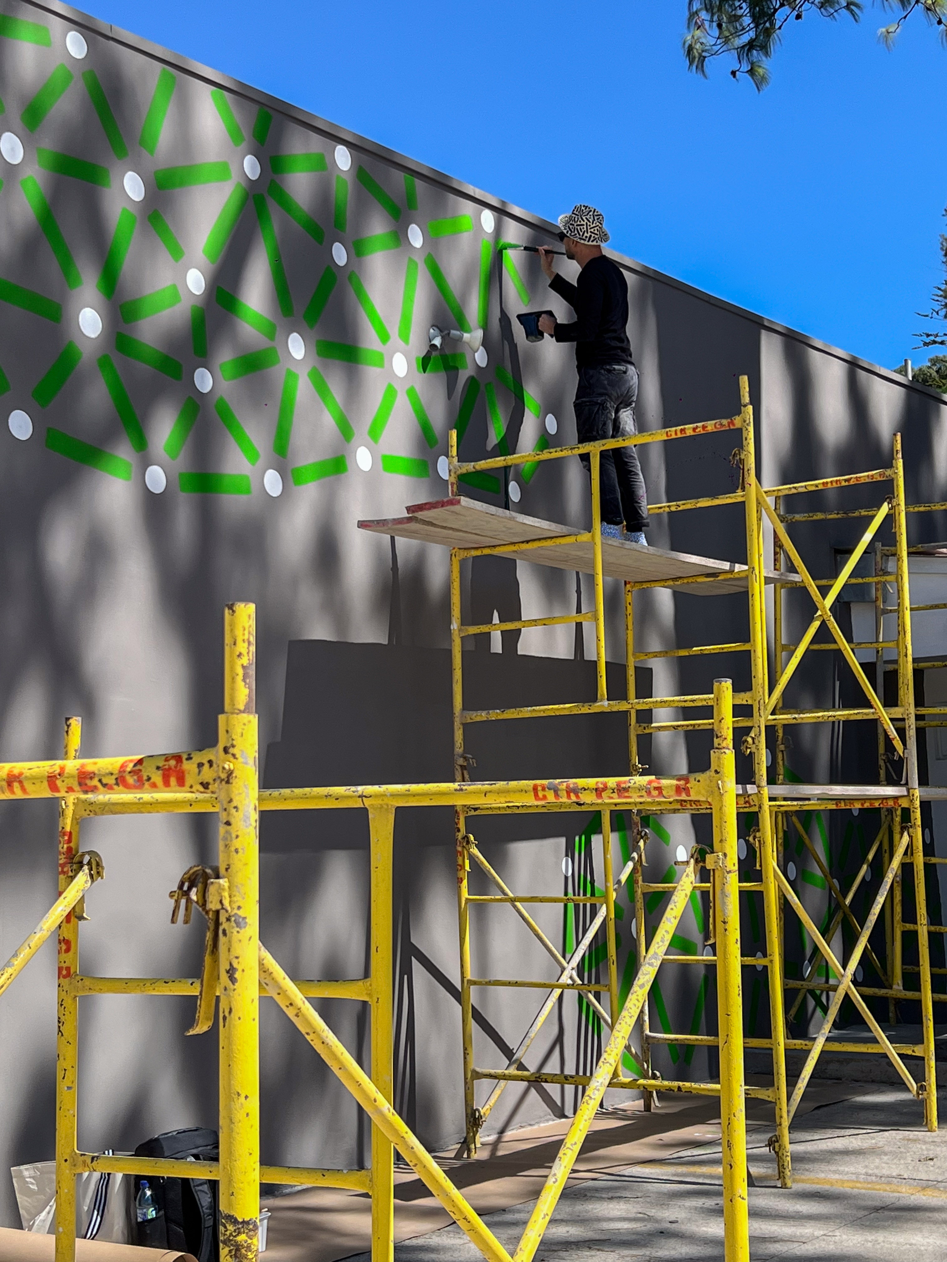 Roots: Artist works from yellow scaffolding to paint green lines and pale dots on a tall exterior wall.